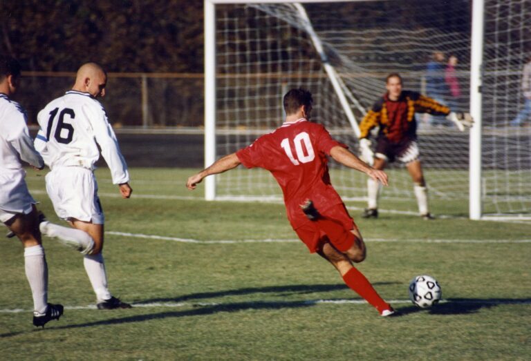 Association football (soccer), Bloomington, Indiana. The offensive player (in red) is Chris Klein of the Indiana Hoosiers. He has sprinted past two defenders (in white) and is about to shoot the ball at the goal or pass it across the field to a teammate in front of the goal.