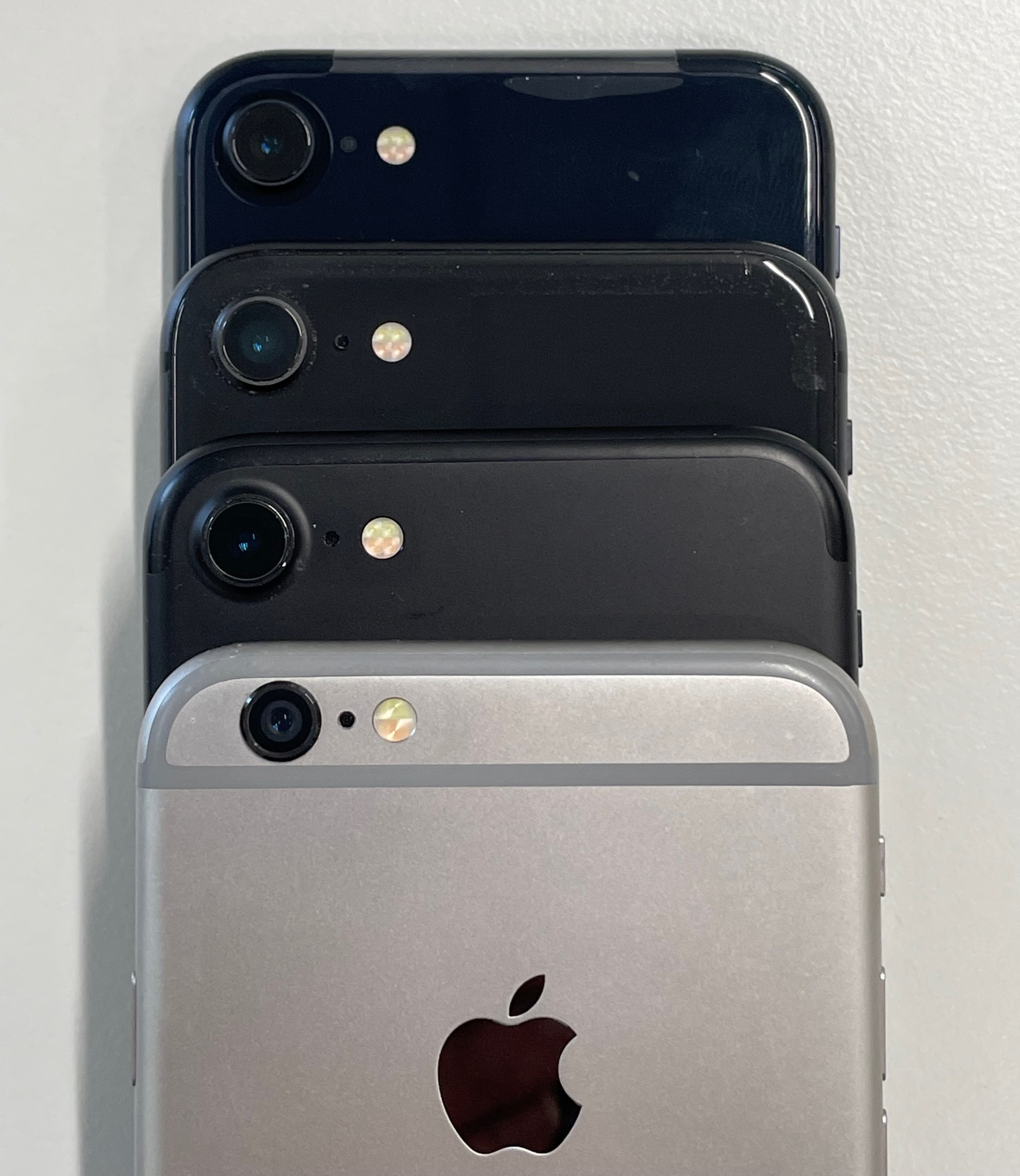 Iphones - models 6, 7, 8 and SE2 - stacked for comparison. These models have nearly identical case shape and dimensions. View of the back side top section, showing: camera, microphone hole, dual-tone LED illuminator. 6 and 7 have aluminium backs whilst 8 and SE have glass. iPhone 6 is "space grey" colour, others are black or "midnight". The SE 2nd gen is new out of box and still has protective shipping wrapper attached; others are used and showing some wear.